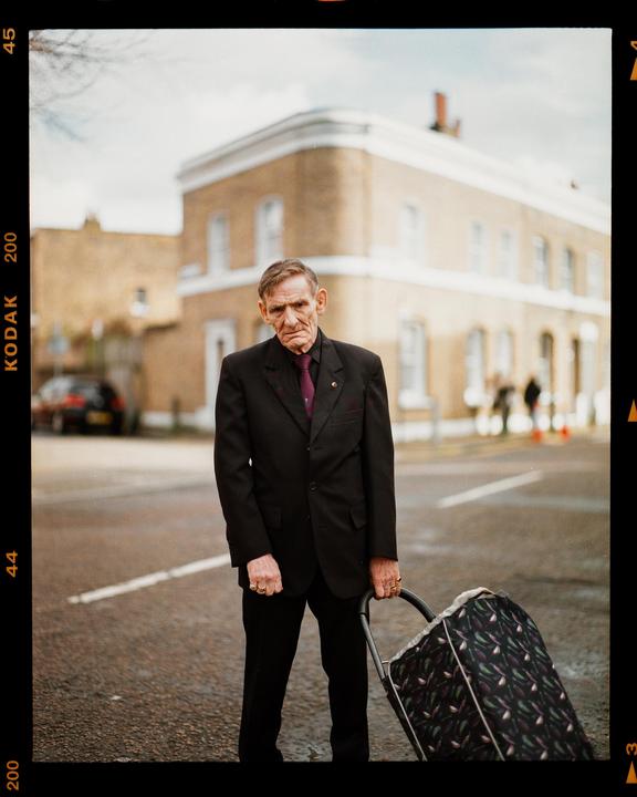 A man in a suit stands on a street holding a shopping trolley.