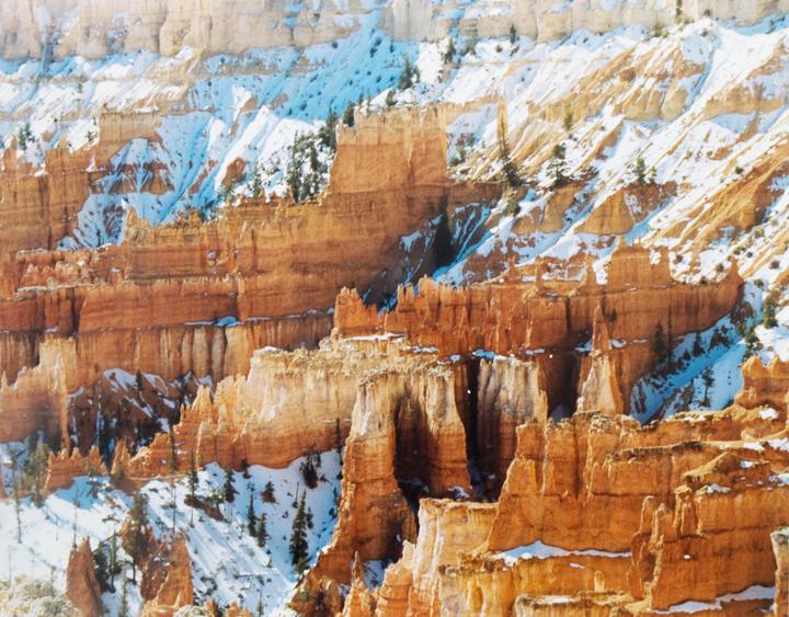 A vast canyon landscape with orange rock formations is partially covered in snow.
