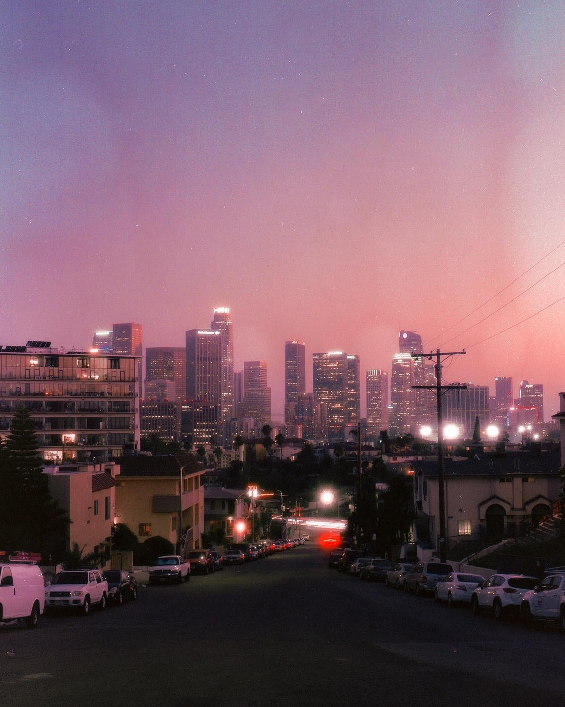 A city skyline with illuminated buildings is visible over a street lined with parked cars at dusk.