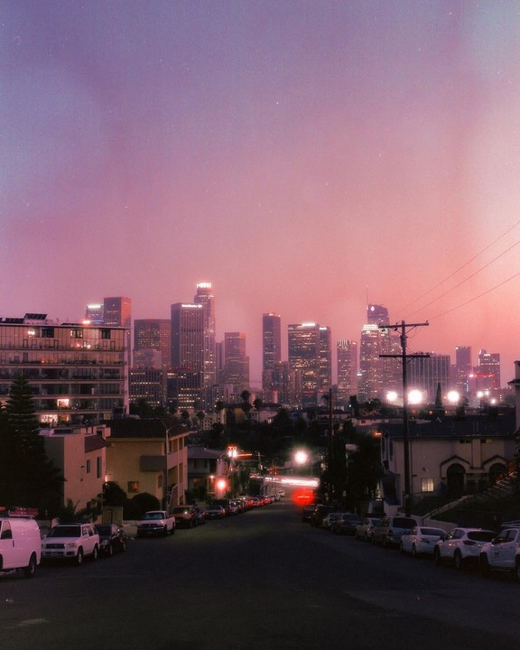 A city skyline with illuminated buildings is visible over a street lined with parked cars at dusk.