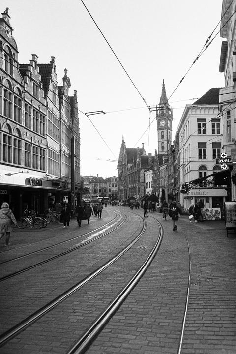 A street with tram tracks and people walking is lined with buildings, including a prominent clock tower in the distance.