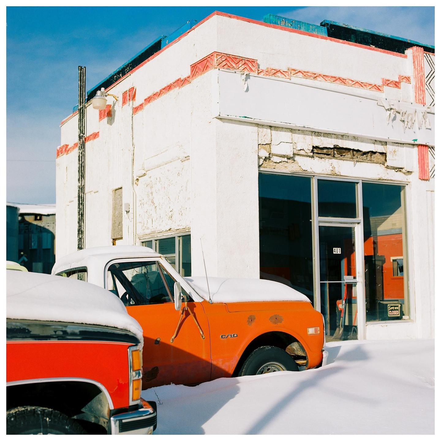 Two snow-covered pickup trucks are parked in front of a white building with red and blue trim.