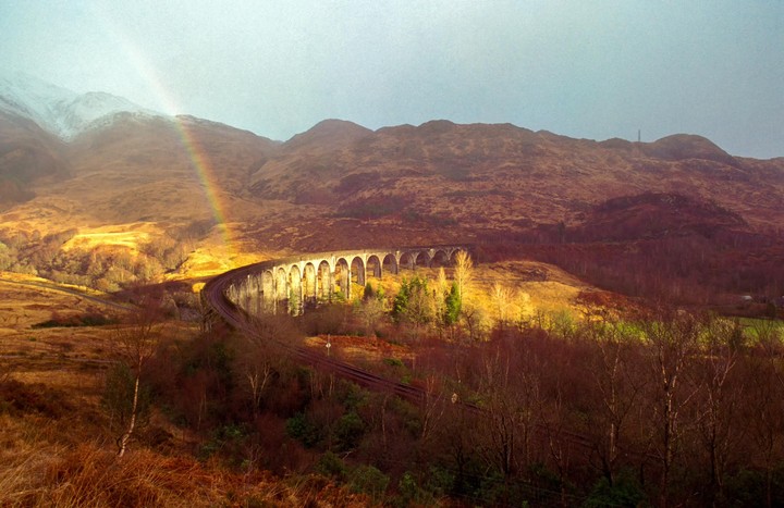A rainbow arches over a stone viaduct with a train track, surrounded by mountains and trees.