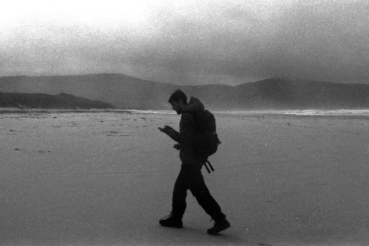 A man with a backpack walks across a wide, flat beach with mountains in the background.
