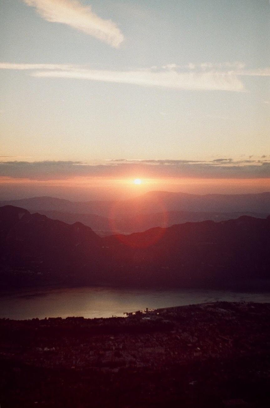 The sun sets over a mountain range and a body of water, with a small town visible on the shore.