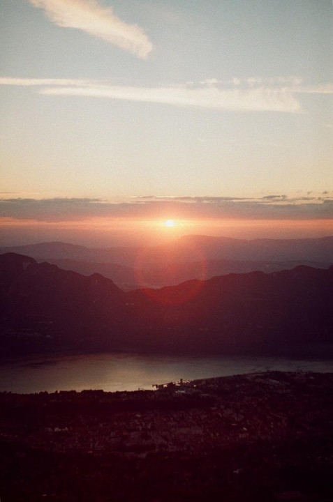 The sun sets over a mountain range and a body of water, with a small town visible on the shore.
