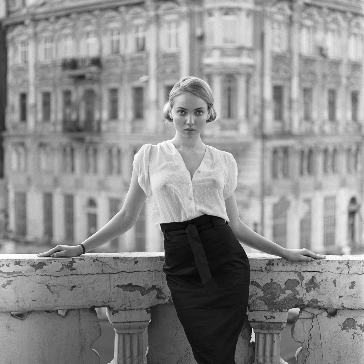 A woman stands on a balcony with a building in the background.