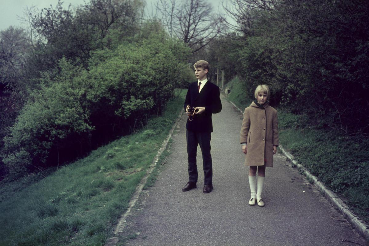 A boy and a girl stand on a paved path surrounded by green foliage.