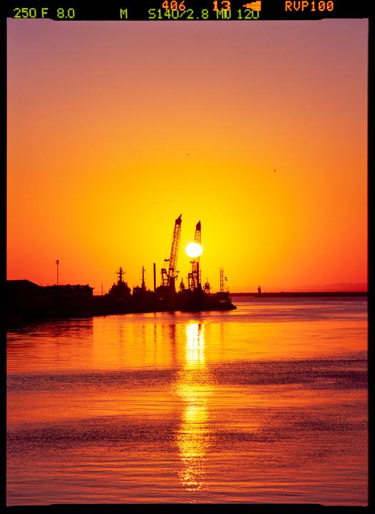 The sun sets behind industrial cranes and ships docked at a harbor, casting an orange glow over the water.