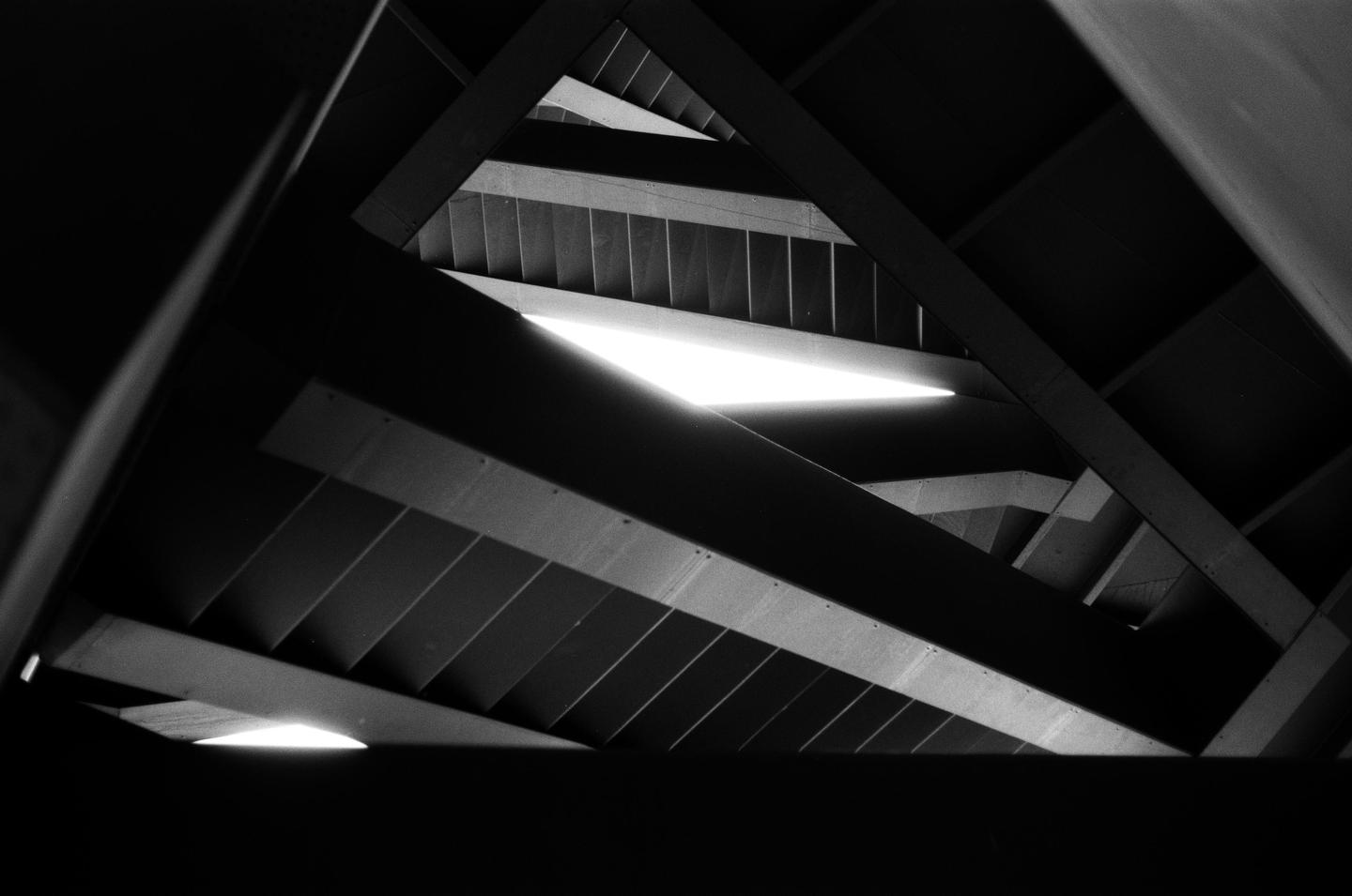 A low-angle, black and white shot shows a complex arrangement of beams and slats, with bright light sources visible through gaps.