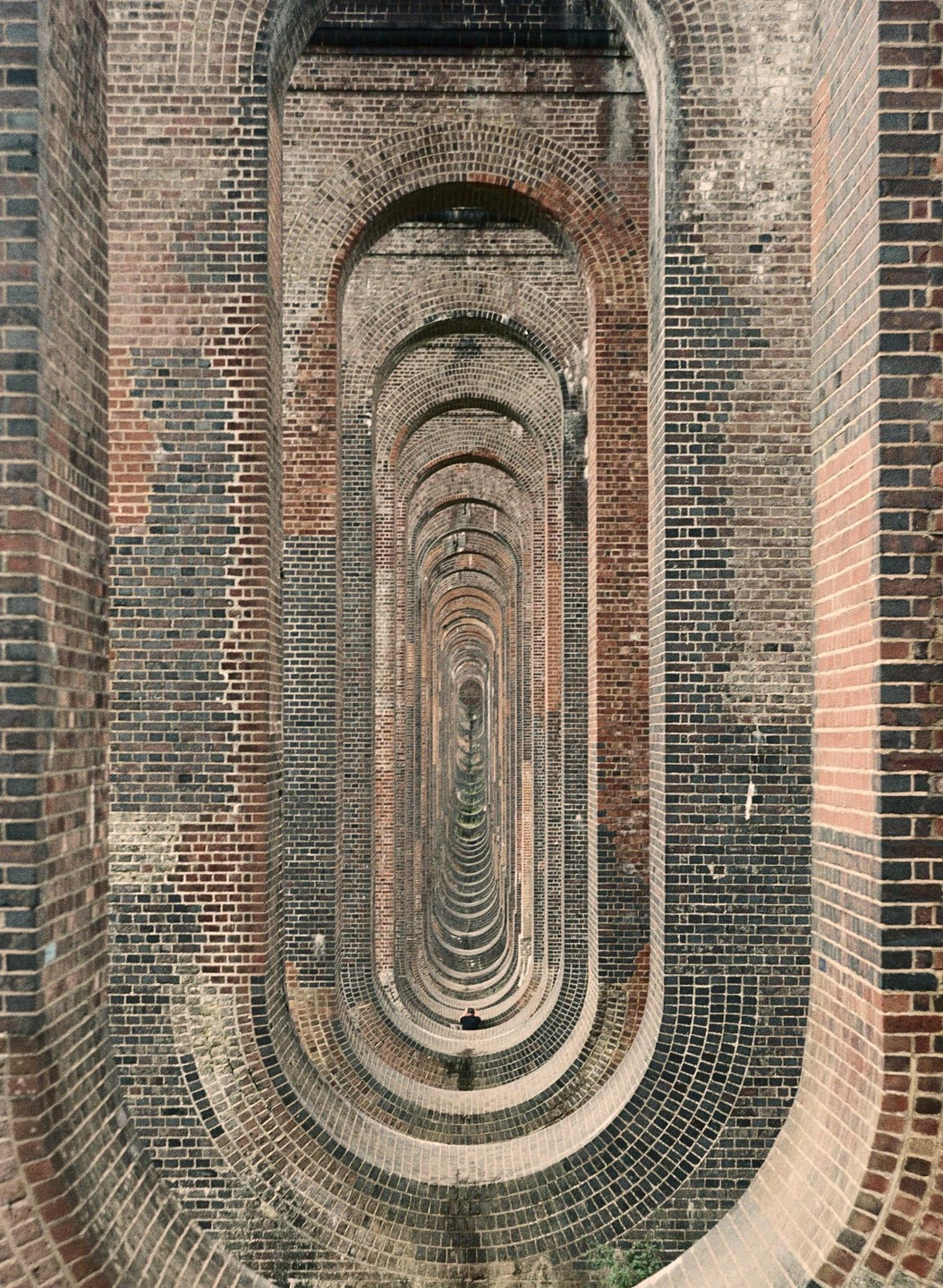 A person sits at the end of a long brick tunnel made of repeating arches.