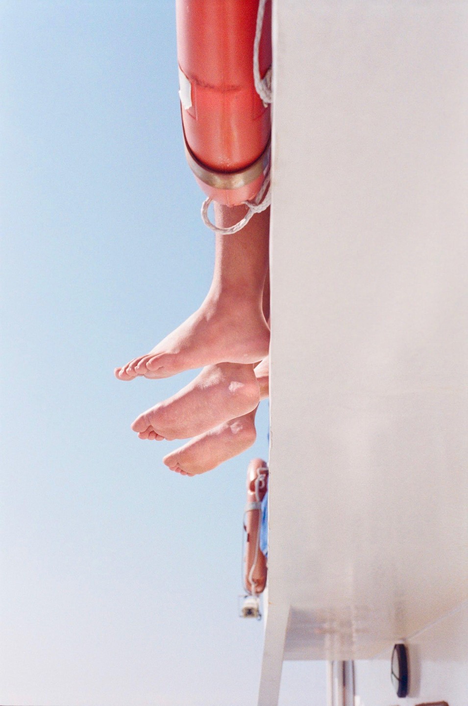 Three pairs of feet dangle over the side of a boat with a life preserver visible against a clear sky.