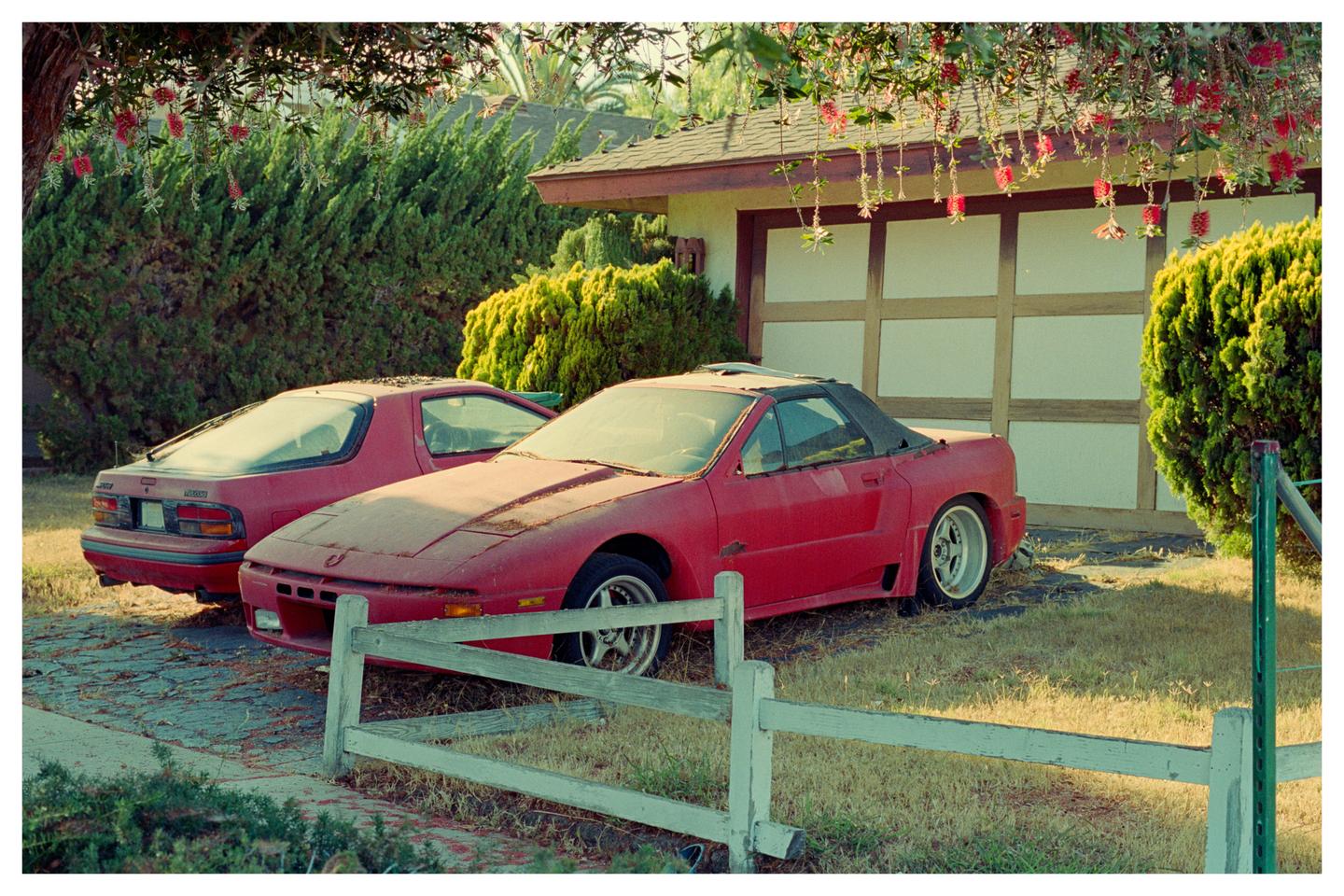 Two red sports cars are parked in a driveway next to a garage, with a white fence in the foreground.