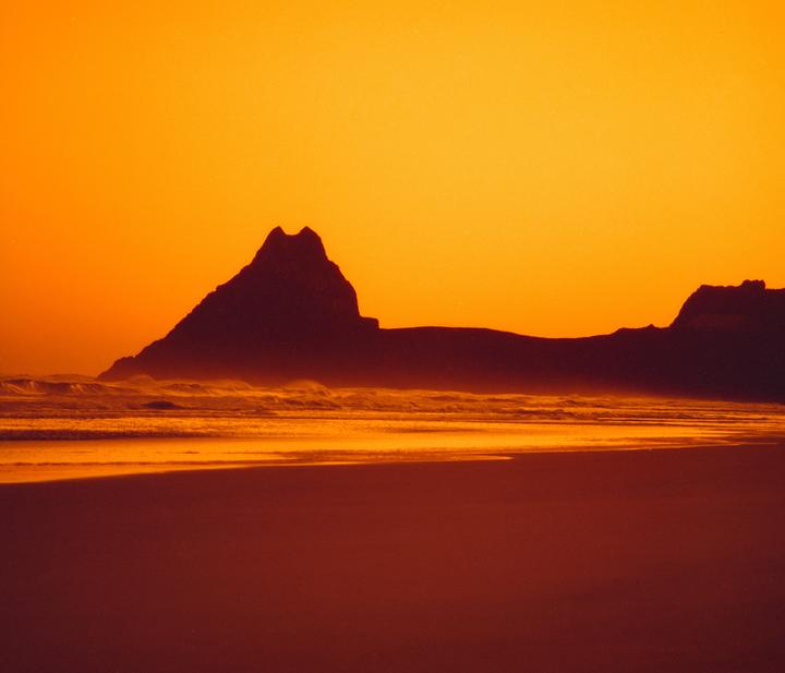 A silhouetted mountain range rises above a beach with waves under an orange sky.