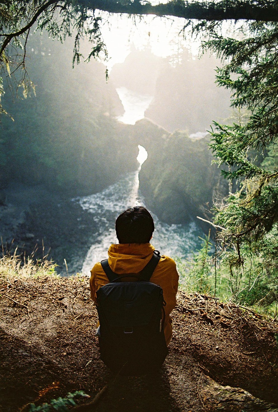 A person wearing a yellow jacket and a backpack sits on a cliff overlooking a river and a bright, hazy landscape.
