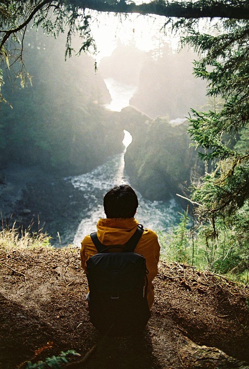 A person wearing a yellow jacket and a backpack sits on a cliff overlooking a river and a bright, hazy landscape.