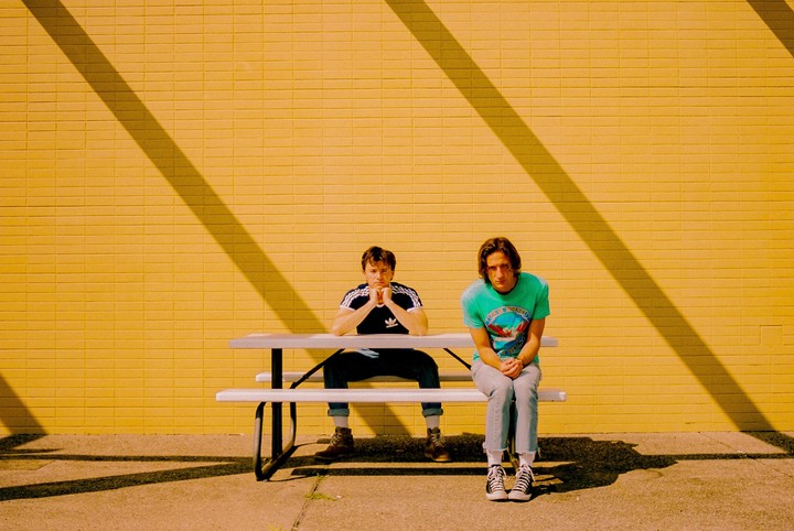Two men sit on a picnic bench in front of a yellow brick wall with a diagonal shadow.