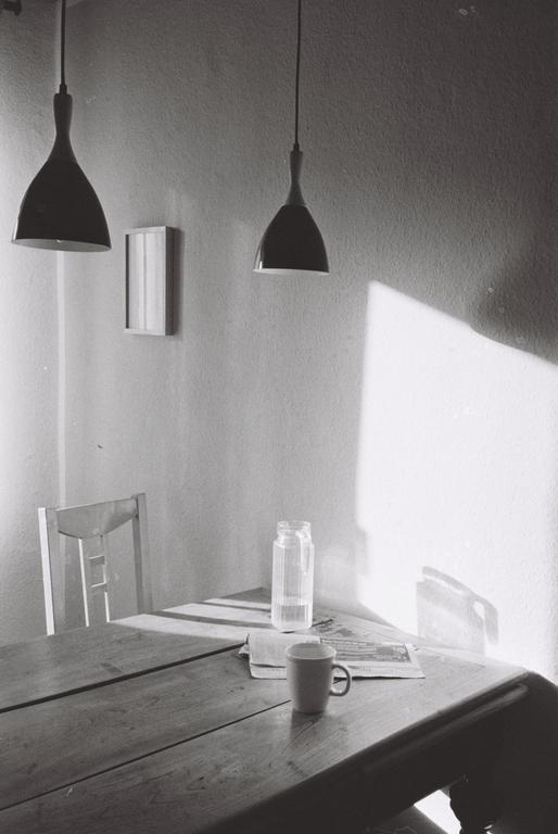 Sunlight streams across a wooden table set with a mug, newspaper, and glass jar, beneath two hanging pendant lamps.