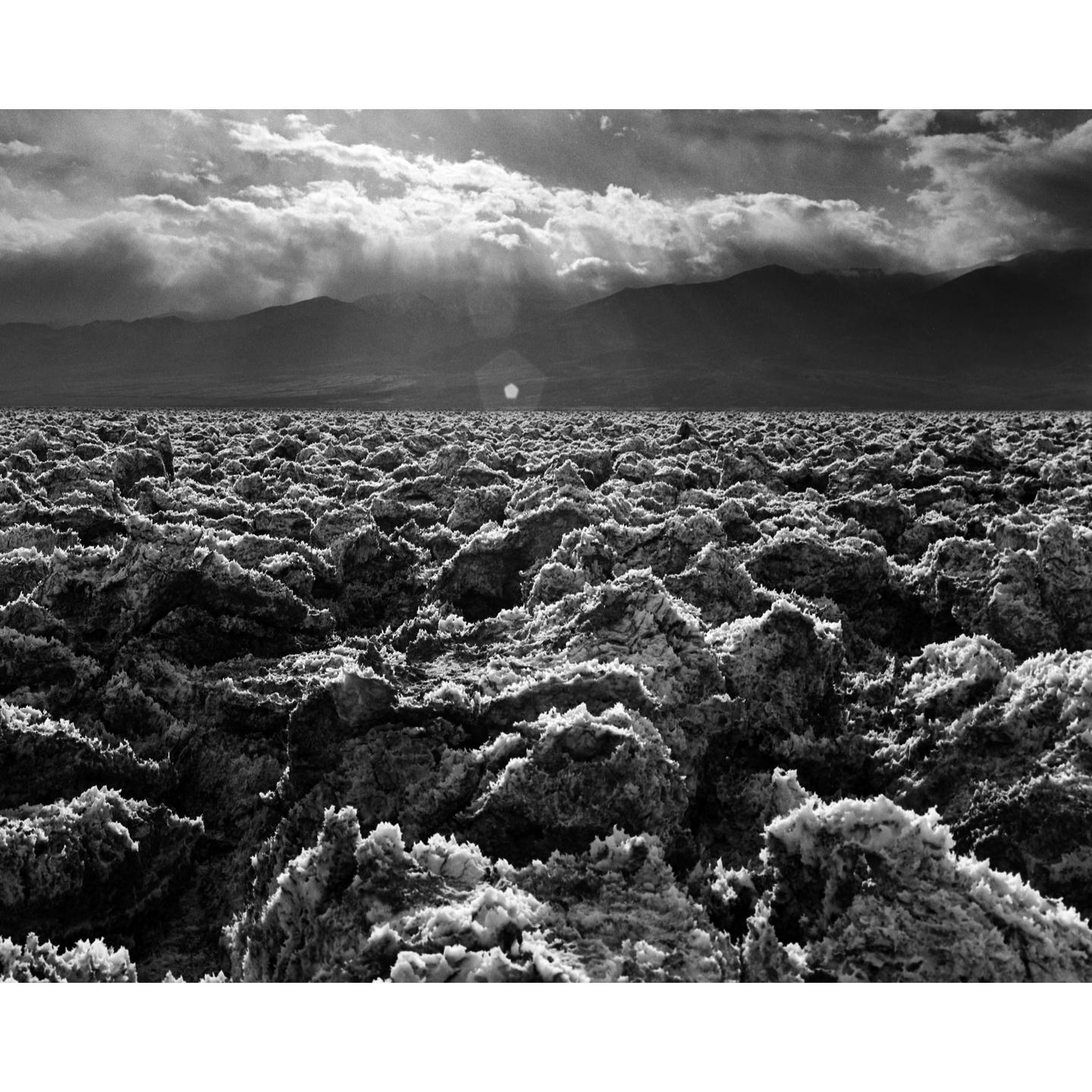 A vast, rugged landscape of salt formations stretches towards distant mountains under a cloudy sky with sun rays.