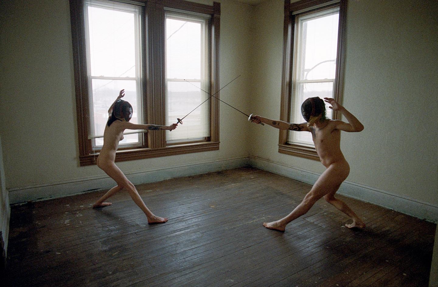 Two nude people wearing fencing masks and holding foils are engaged in a duel in an empty room with three windows.