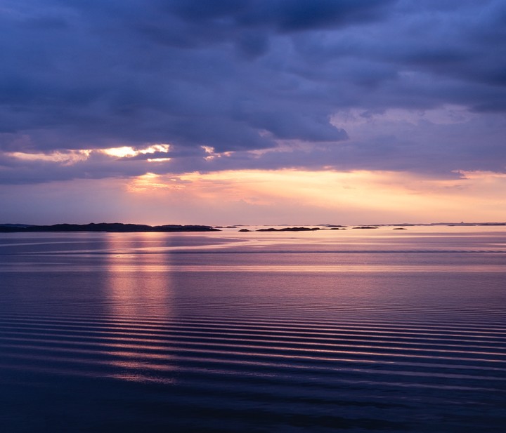 The sun sets over a body of water with small islands in the distance and ripples on the surface.