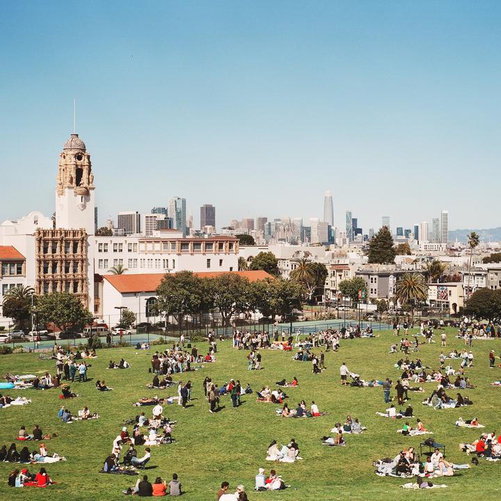 A large crowd of people are relaxing on a grassy hill overlooking a city skyline with a prominent tower in the foreground.