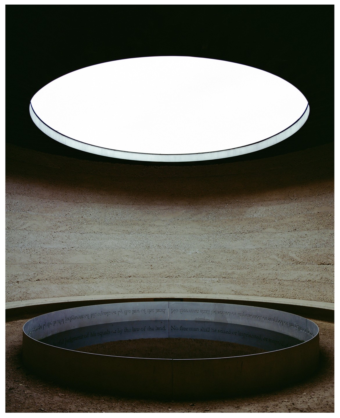 A circular skylight illuminates a circular metal band with text on a textured wall and ground.