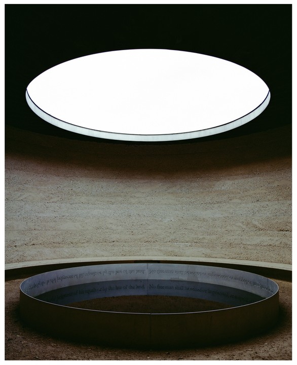 A circular skylight illuminates a circular metal band with text on a textured wall and ground.