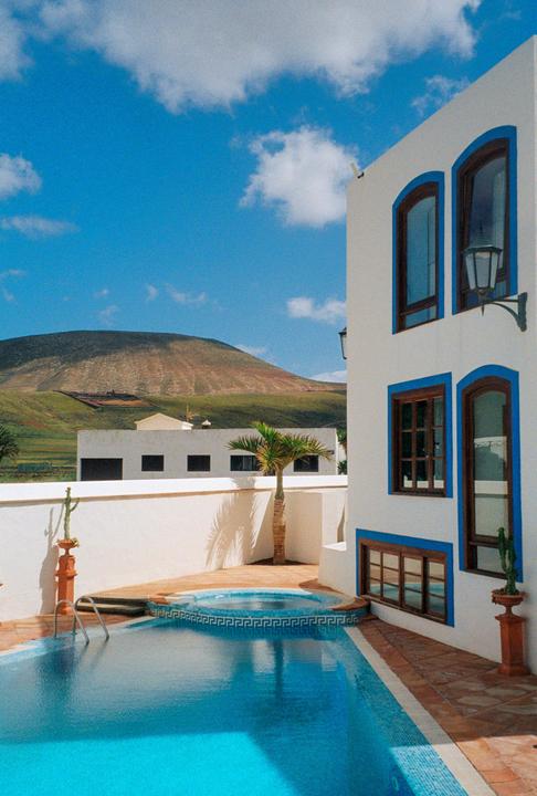 A white building with blue trim and brown windows stands next to a swimming pool and hot tub, with a mountain in the background.