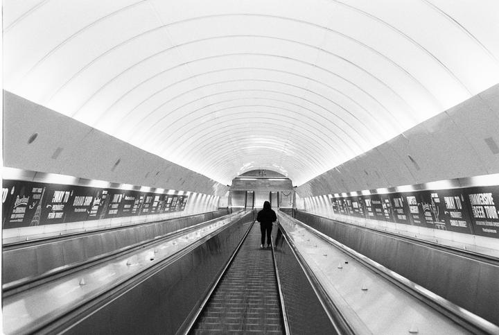 A person stands on a moving walkway in a long, curved tunnel with advertisements on the walls.