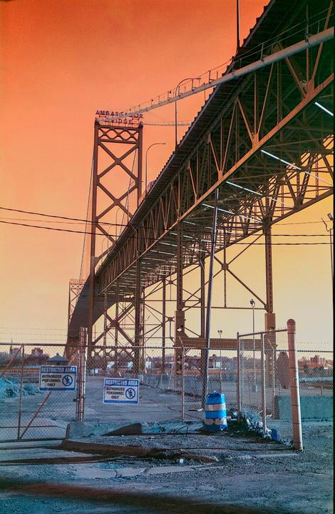 A large metal bridge with a sign that reads "Ambassador Bridge" is visible against an orange sky, with a chain-link fence and two "restricted area" signs in the foreground.