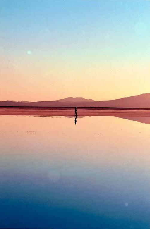 A person stands in a shallow body of water reflecting the colorful sky and distant mountains at sunset.
