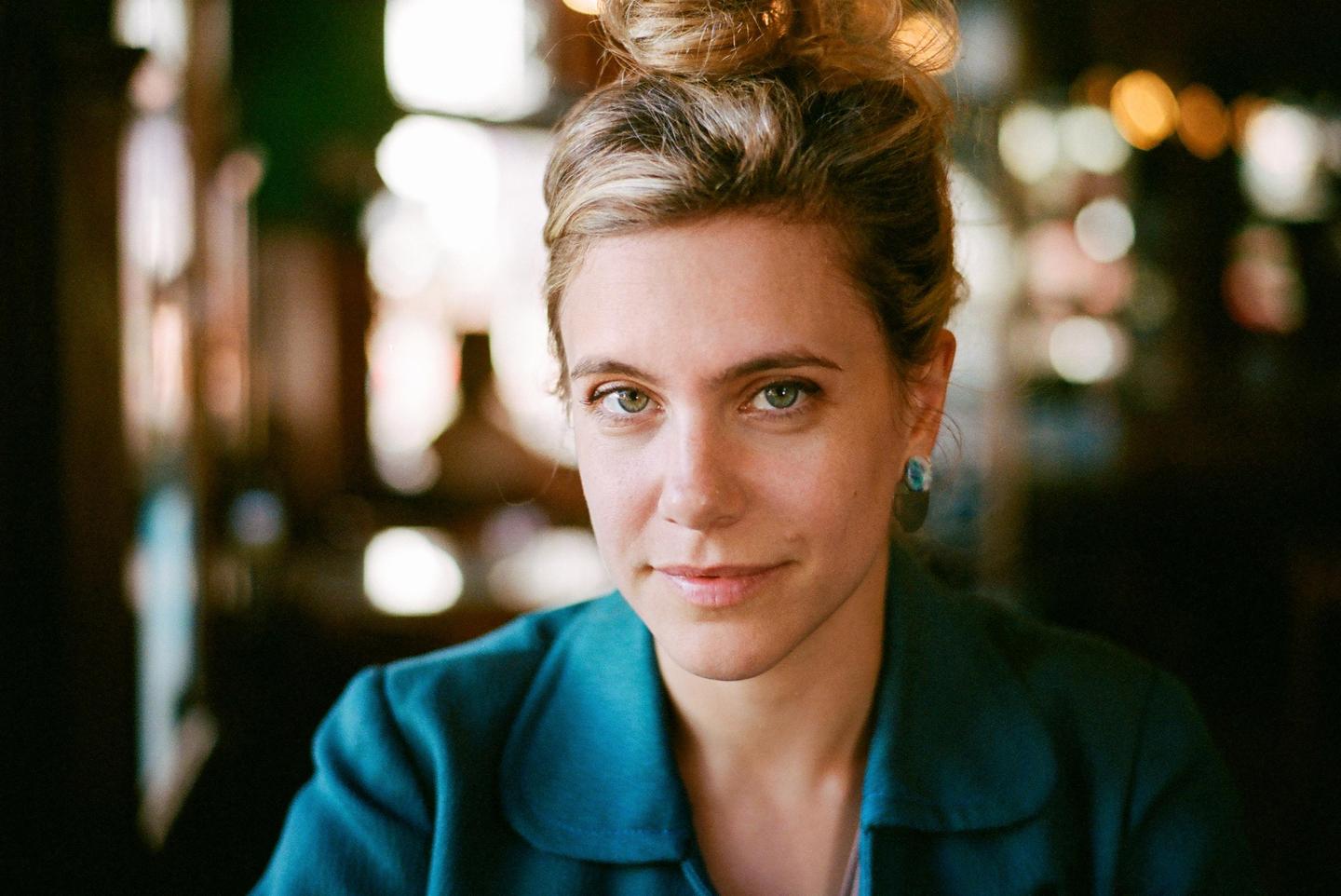 A woman with her hair in a bun and wearing a teal jacket looks directly at the viewer.
