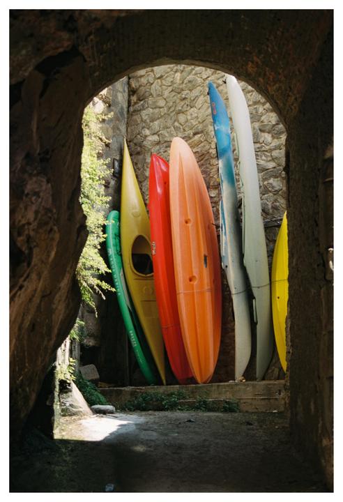 Several colorful kayaks are stored upright against a stone wall, viewed through a stone archway.