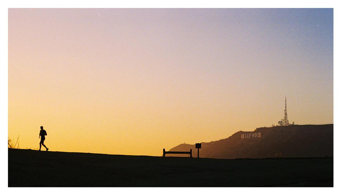 A man runs up a hill with the Hollywood sign and a radio tower visible in the distance at sunset.