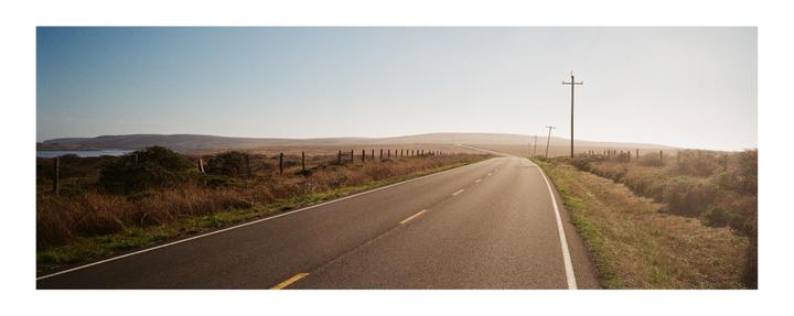 A long, empty road stretches into the distance with hills and a body of water visible under a clear sky.