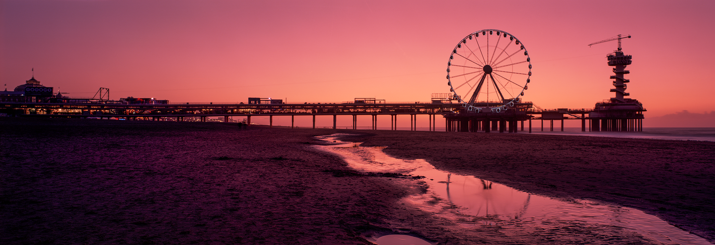 A long pier with a ferris wheel and a tower stands over a beach with a stream of water reflecting the sunset.