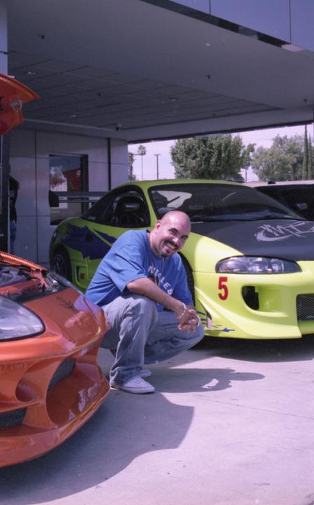 A man crouches next to a bright yellow sports car with an orange sports car in the foreground.