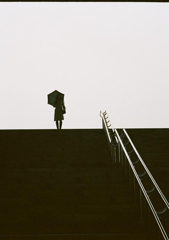 A woman holding an umbrella stands on a dark elevated surface next to a railing.