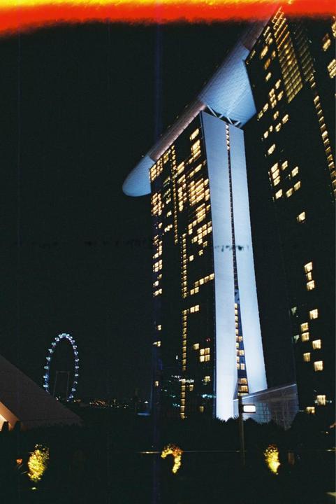 A tall, illuminated building with a boat-like structure on top stands against a dark night sky, with a Ferris wheel visible in the distance.