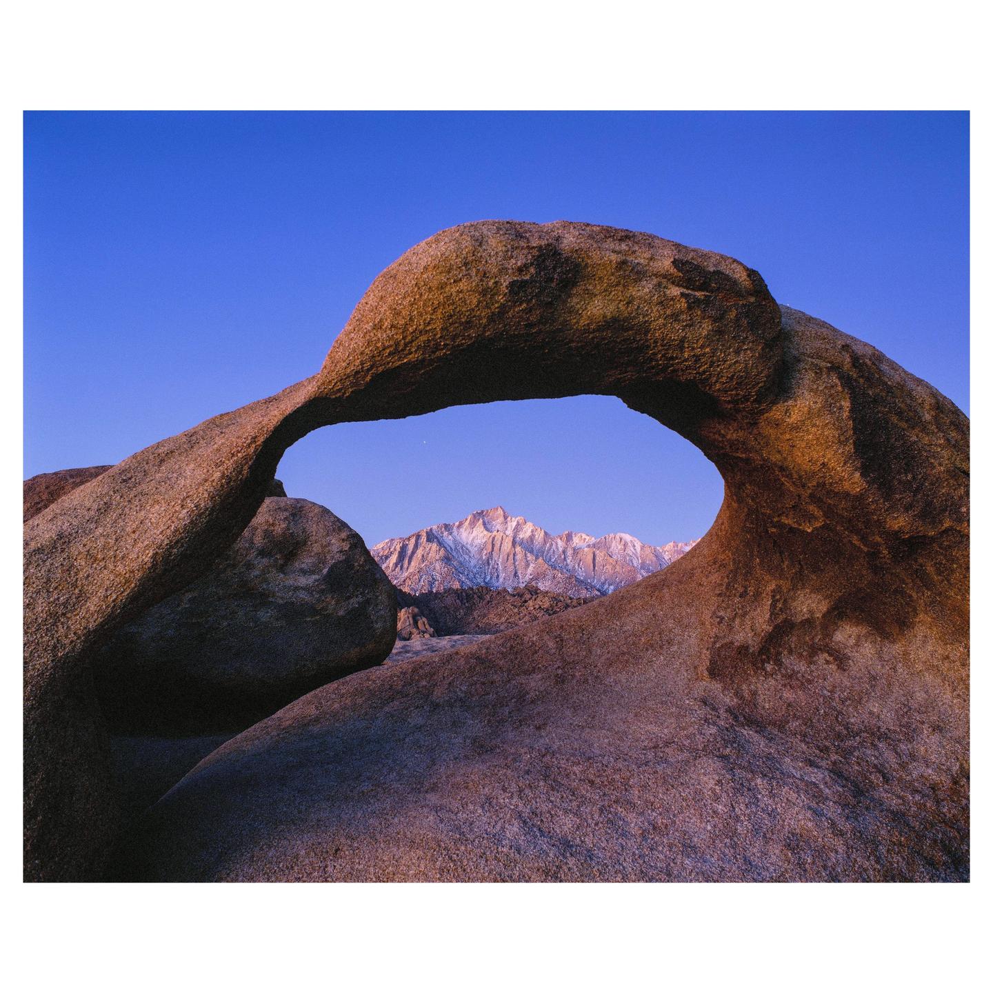 A natural rock arch frames a snow-capped mountain range under a clear blue sky.