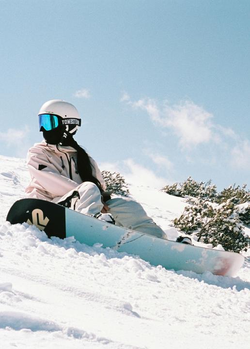 A woman in a white helmet and blue goggles sits on a snowy mountain slope next to a snowboard.