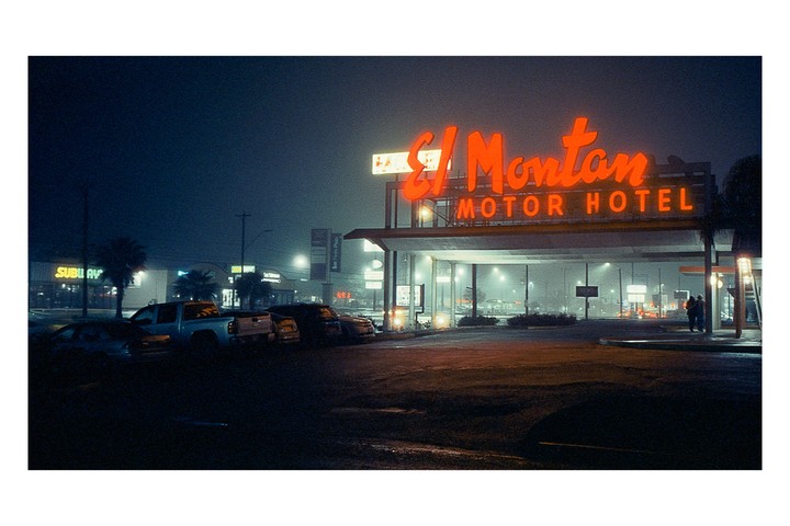 A neon sign for the El Montan Motor Hotel glows orange in the fog at night, with cars parked in the foreground and a Subway restaurant sign visible in the background.