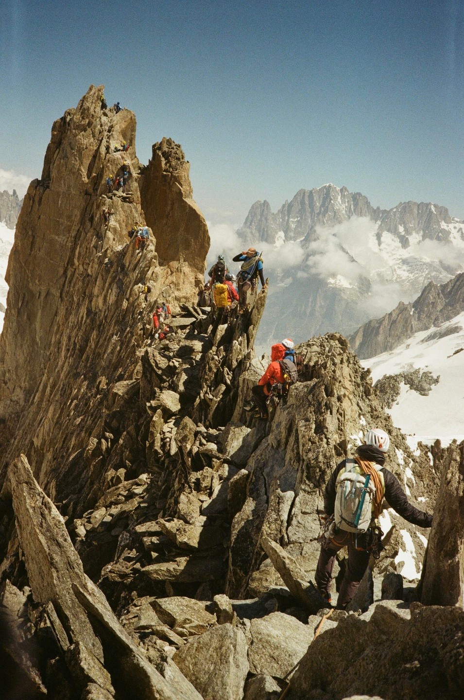 A group of climbers ascends a rocky mountain ridge with snow-capped peaks and clouds in the background.