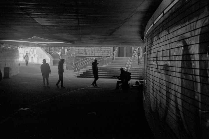 A street musician plays guitar under a bridge while people walk by.