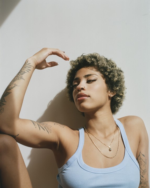 A woman with curly hair and tattoos poses against a white wall with her arm raised.
