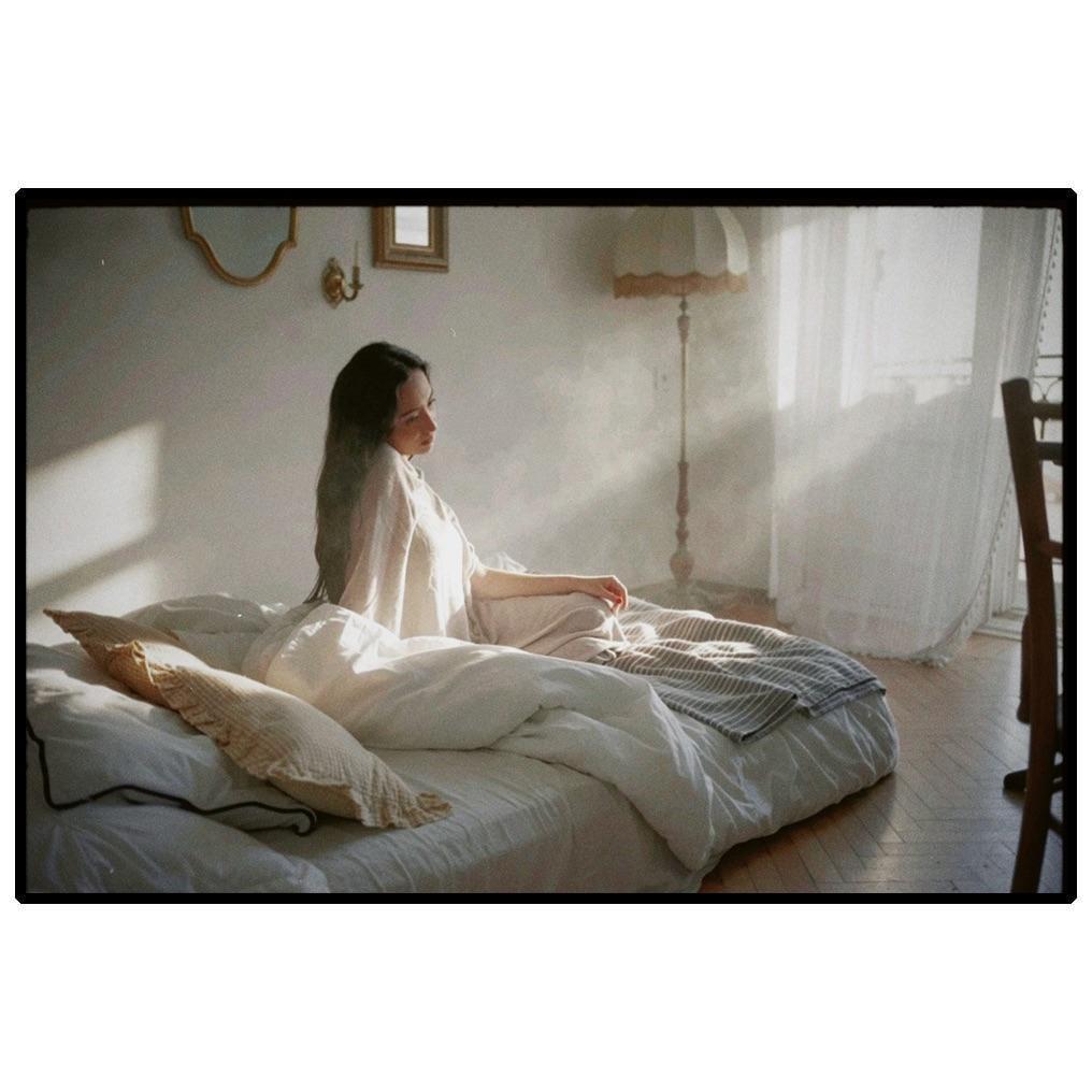 A woman sits on a bed in a sunlit room.