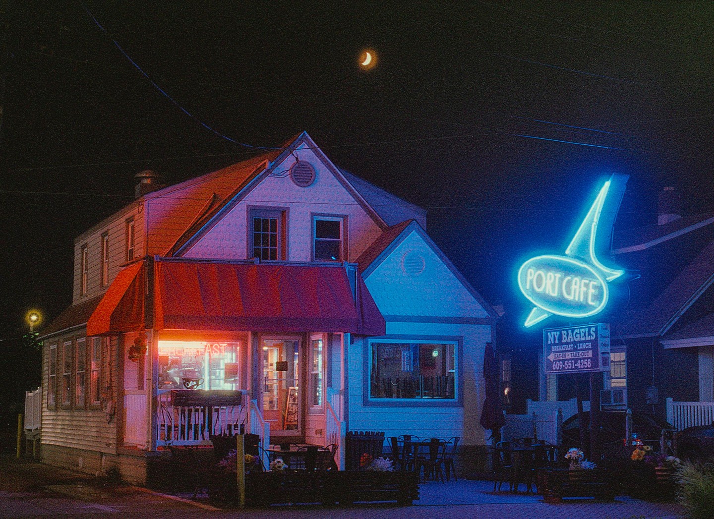 A cafe with a red awning and a blue neon sign shaped like a fish is illuminated at night under a crescent moon.