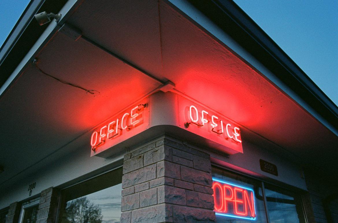 Two red neon "OFFICE" signs are visible on the corner of a building with a blue neon "OPEN" sign below them.