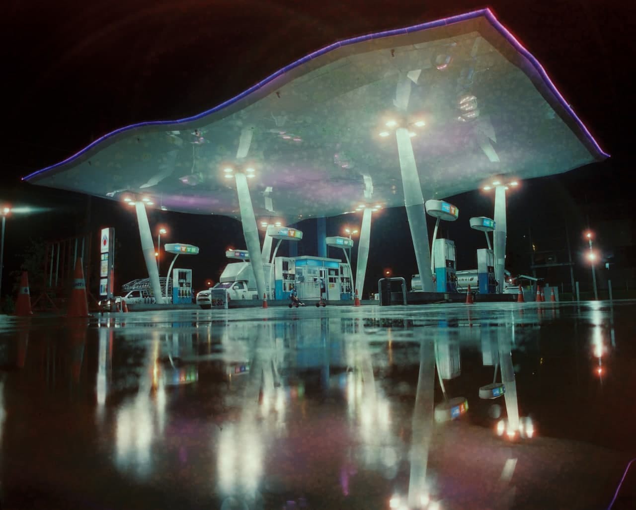 A gas station with a large, illuminated canopy is reflected in a wet ground at night.